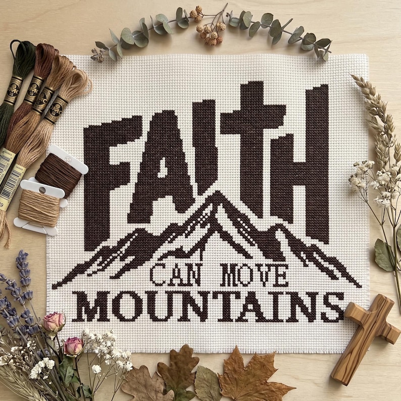 May include: A cross-stitch embroidery on white Aida fabric featuring the dark brown text FAITH CAN MOVE MOUNTAINS with a mountain range graphic. The piece is surrounded by embroidery floss skeins, dried lavender, eucalyptus, and a wooden cross.