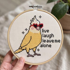 K&ouml;nnte beinhalten: Ein runder Stickrahmen aus Holz mit einem Kreuzstichmotiv eines gelben Nymphensittichs, der eine rote herzf&ouml;rmige Sonnenbrille tr&auml;gt. Der Vogel hat rosa Wangen und ist von kleinen schwarzen Sternen umgeben. Der Text lautet: live laugh leave me alone.