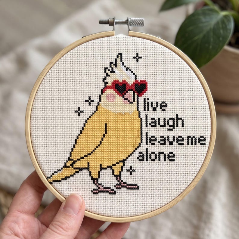 K&ouml;nnte beinhalten: Ein runder Stickrahmen aus Holz mit einem Kreuzstichmotiv eines gelben Nymphensittichs, der eine rote herzf&ouml;rmige Sonnenbrille tr&auml;gt. Der Vogel hat rosa Wangen und ist von kleinen schwarzen Sternen umgeben. Der Text lautet: live laugh leave me alone.