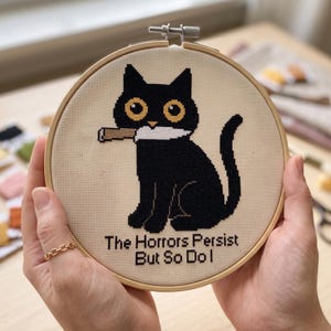 May include: A circular wooden embroidery hoop containing a cross-stitch design of a black cat with large yellow eyes holding a white knife in its mouth. Below the cat, the text reads, "The Horrors Persist But So Do I" in black stitched letters.