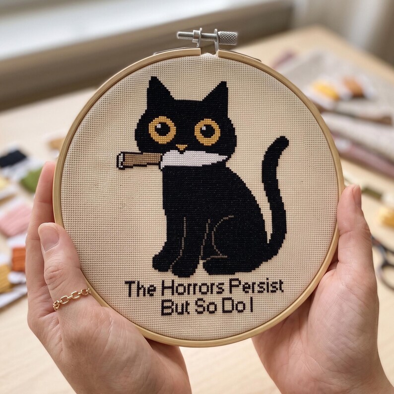 May include: A circular wooden embroidery hoop containing a cross-stitch design of a black cat with large yellow eyes holding a white knife in its mouth. Below the cat, the text reads, "The Horrors Persist But So Do I" in black stitched letters.