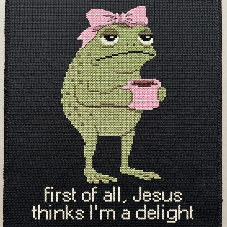 Op de afbeelding: Een kruissteekborduurwerk op zwarte stof met een groene pad die een roze strik draagt en een roze mok vasthoudt. Onder de pad staat de tekst: First of all, Jesus thinks I'm a delight. De pad heeft een neutrale uitdrukking en textuur.