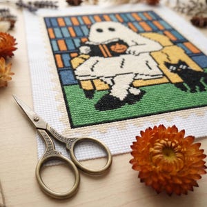 K&ouml;nnte beinhalten: Ein Kreuzstichdesign mit einem Geist, der ein Buch mit einem K&uuml;rbis und einer schwarzen Katze liest. Das Design ist mit einem wei&szlig;en Rand versehen. Eine goldene Schere und orangefarbene Blumen befinden sich im Vordergrund.