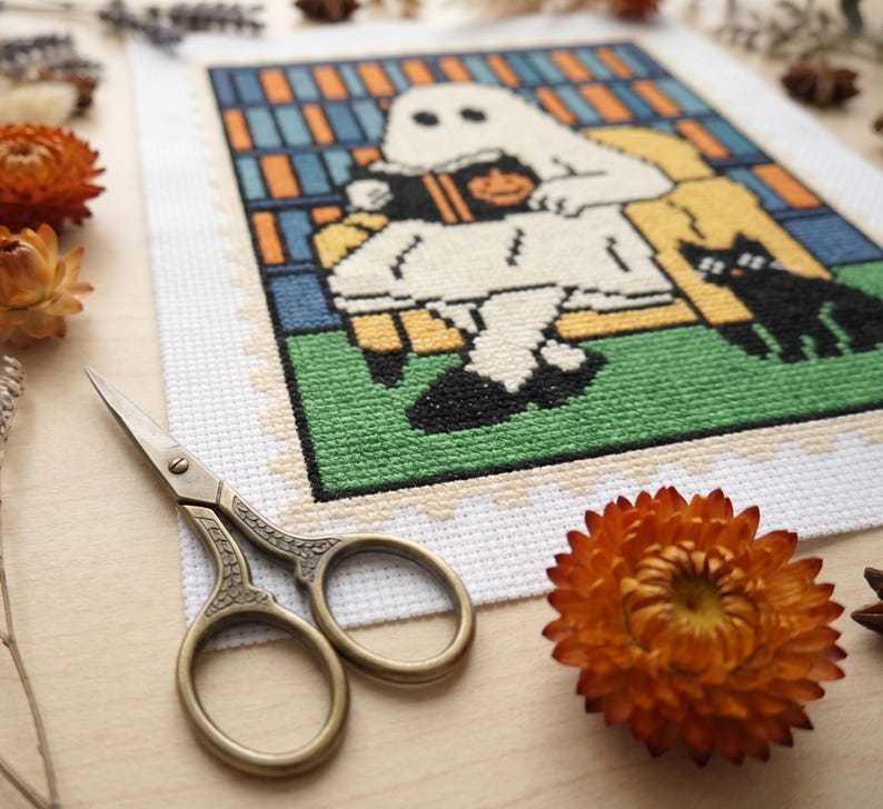 K&ouml;nnte beinhalten: Ein Kreuzstichdesign mit einem Geist, der ein Buch mit einem K&uuml;rbis und einer schwarzen Katze liest. Das Design ist mit einem wei&szlig;en Rand versehen. Eine goldene Schere und orangefarbene Blumen befinden sich im Vordergrund.