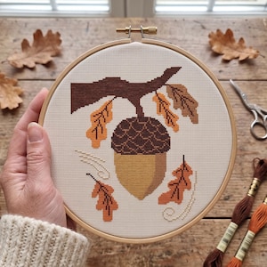 May include: A circular wooden embroidery hoop containing a cross-stitch pattern of a large brown acorn hanging from a dark brown oak branch. The design features several small orange and brown oak leaves surrounding the acorn on white fabric.