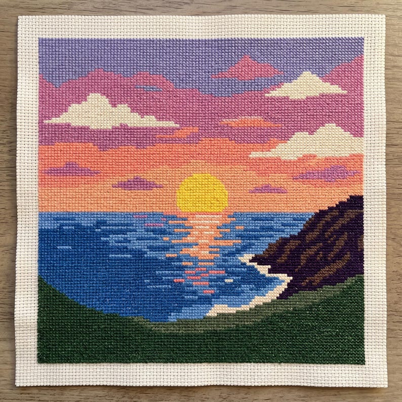 May include: A square cross-stitch embroidery depicting a sunset over the ocean. The scene features a bright yellow sun, a blue sea with orange reflections, a dark purple cliffside, and a green foreground under a pink, purple, and orange sky.