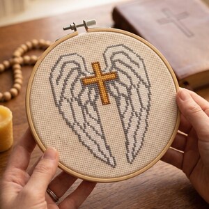 May include: A circular wooden embroidery hoop containing a cross-stitch design on off-white fabric. The design features a pair of large, detailed white and grey wings centered behind a small, solid gold-colored cross with a textured finish.