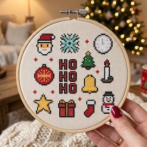 Op de afbeelding: Een ronde houten borduurring met een kruissteekpatroon op beige stof. Het ontwerp bevat kersticonen zoals de Kerstman, een sneeuwvlok, een klok, een boom, een kaars, een bel, een ster, een cadeau, een kerstsok en een sneeuwpop, met de tekst HO HO HO.