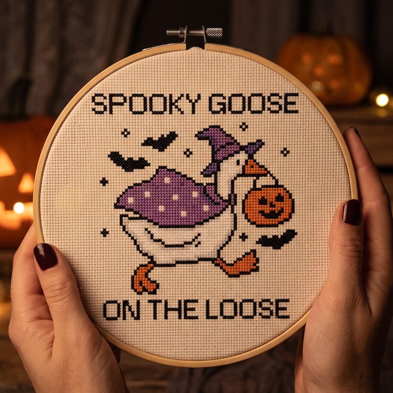 May include: A circular wooden embroidery hoop containing a cross-stitch design on beige fabric. The design features a white goose wearing a purple witch hat and cape, carrying an orange jack-o'-lantern. Text reads: SPOOKY GOOSE ON THE LOOSE.