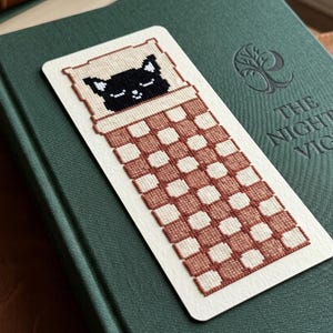 Pode incluir: Um marcador de livros retangular com um gato preto pixelizado dormindo em uma cama com um padr&atilde;o xadrez marrom e branco. O marcador de livros est&aacute; em um livro verde com o t&iacute;tulo "THE NIGHT'S VIGIL".