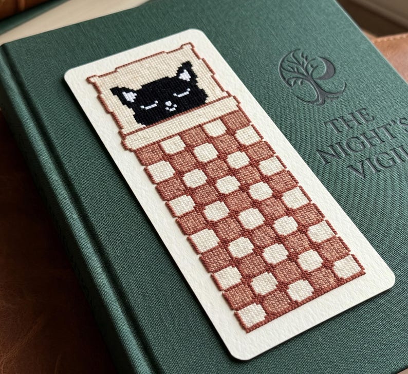 Pode incluir: Um marcador de livros retangular com um gato preto pixelizado dormindo em uma cama com um padr&atilde;o xadrez marrom e branco. O marcador de livros est&aacute; em um livro verde com o t&iacute;tulo "THE NIGHT'S VIGIL".