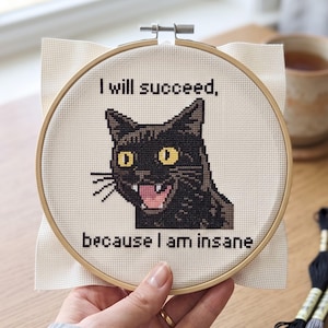 May include: A circular wooden embroidery hoop containing a piece of white Aida fabric with a cross-stitch design of a black cat with yellow eyes and an open mouth. The text reads: I will succeed, because I am insane in black lettering.