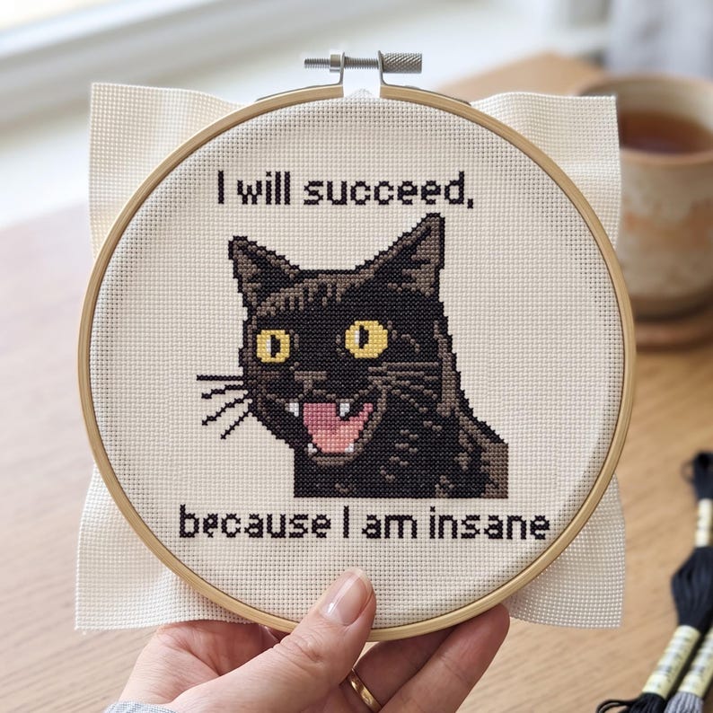 May include: A circular wooden embroidery hoop containing a piece of white Aida fabric with a cross-stitch design of a black cat with yellow eyes and an open mouth. The text reads: I will succeed, because I am insane in black lettering.