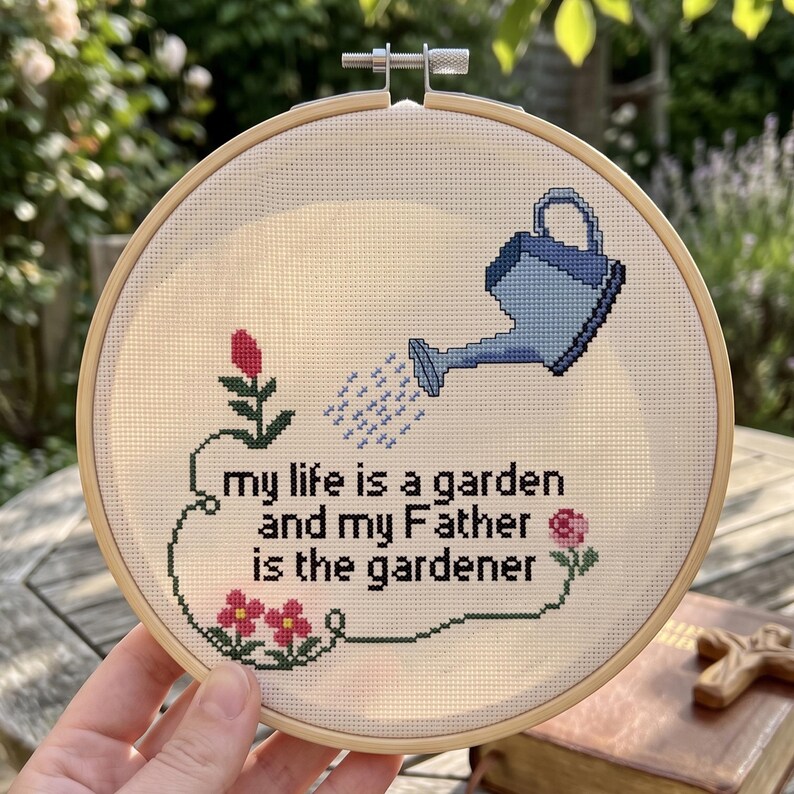Op de afbeelding: Een ronde houten borduurring met witte Aida-stof en een kruissteekontwerp. Het ontwerp toont een blauwe gieter, roze bloemen, groene stengels en de zwarte tekst: my life is a garden and my Father is the gardener.