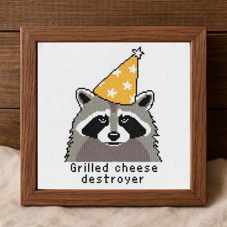 Op de afbeelding: Kruissteekkunst van een wasbeer met een gele feesthoed met witte sterren. De wasbeer heeft zwarte markeringen rond ogen en neus. De tekst "Grilled cheese destroyer" staat eronder, alles in een bruine houten lijst.