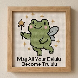 May include: Cross-stitch art in a wooden frame featuring a green frog with fairy wings and a star wand. The frog has pink cheeks and is surrounded by gold stars. Text reads "May All Your Delulu Become Trululu."