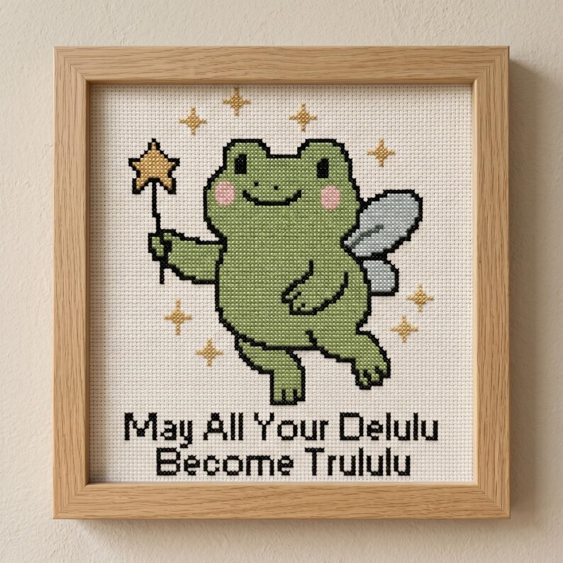 May include: Cross-stitch art in a wooden frame featuring a green frog with fairy wings and a star wand. The frog has pink cheeks and is surrounded by gold stars. Text reads "May All Your Delulu Become Trululu."