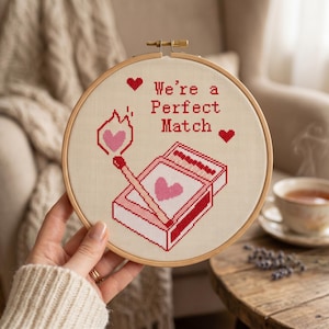Puede incluir: Un bastidor de bordado circular de madera con un dise&ntilde;o de punto de cruz sobre tela color crema. El dise&ntilde;o muestra una caja de cerillas y una cerilla encendida con una llama en forma de coraz&oacute;n rosa, junto al texto rojo We're a Perfect Match.
