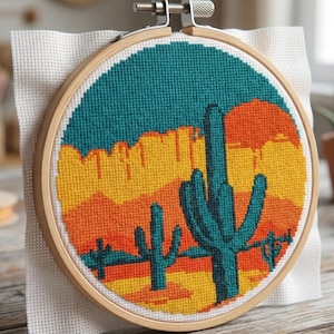 May include: A cross-stitch embroidery hoop featuring a desert landscape with cacti, mountains, and a setting sun. The design uses teal, orange, yellow, and brown thread on white fabric, set in a wooden hoop.