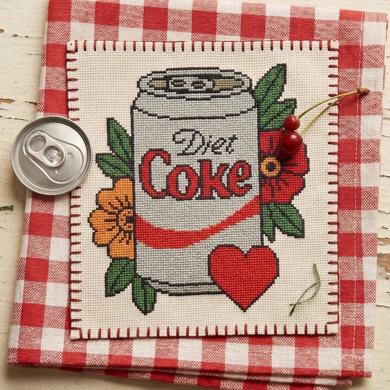 May include: A cross-stitch embroidery on cream fabric featuring a silver Diet Coke can with red lettering, surrounded by orange and red flowers with green leaves and a red heart. A metal soda can lid and two cherries are placed on the fabric.