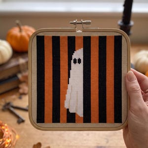 May include: A square wooden embroidery hoop containing a cross-stitch pattern of a white ghost centred against a background of vertical black and orange stripes on a light beige fabric. The ghost features two small black rectangular eyes.