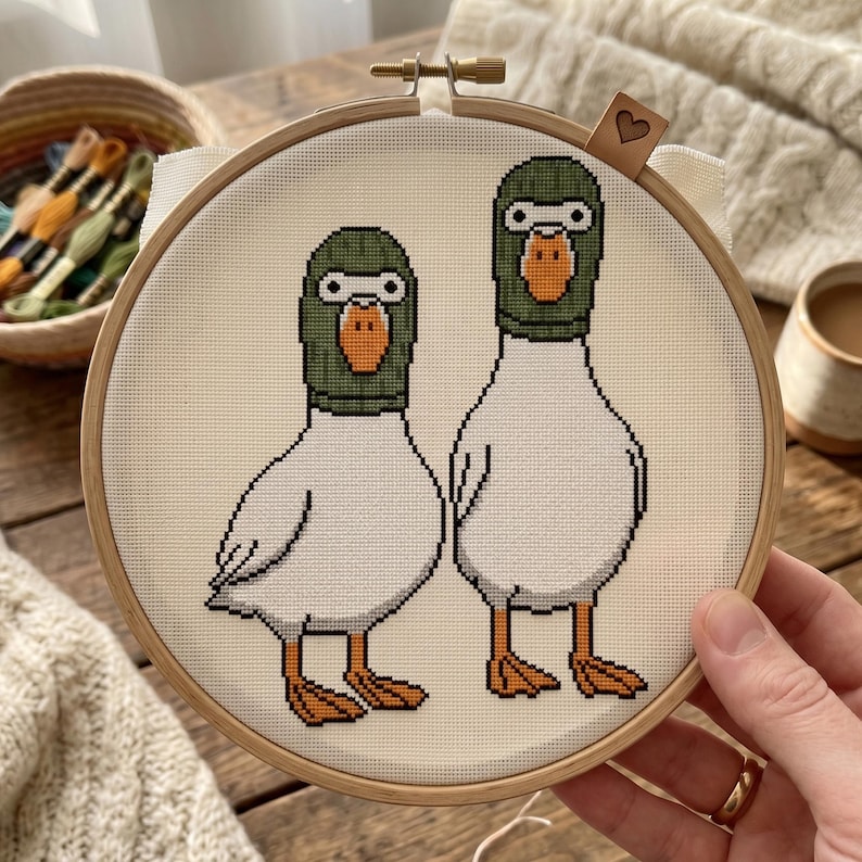 Peut inclure: Un tambour &agrave; broder circulaire en bois contenant un motif au point de croix repr&eacute;sentant deux canards blancs portant des cagoules vertes. Les canards ont des becs et des pattes orange, sur un tissu cr&egrave;me. Une &eacute;tiquette en cuir avec un c&oelig;ur.