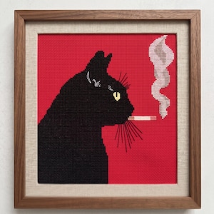 Puede incluir: Arte de punto de cruz de un gato negro fumando un cigarrillo, sobre un fondo rojo. El perfil del gato está detallado con ojos amarillos y largos bigotes. La obra de arte está enmarcada en un marco de madera con un paspartú de lino neutro.