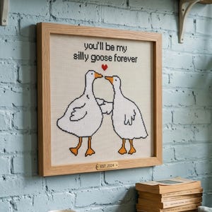 May include: Cross-stitch artwork in a light wood frame. The artwork features two white geese with orange feet and beaks, touching beaks under a red heart. Text above reads "you'll be my silly goose forever". The bottom reads "EST. 2024".