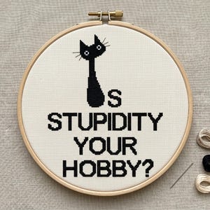 Pu&ograve; includere: Un telaio da ricamo circolare in legno contenente tessuto bianco con un disegno a punto croce nero raffigurante la sagoma di un gatto e il testo IS STUPIDITY YOUR HOBBY? Il disegno &egrave; centrato sul tessuto, teso da una vite in metallo.
