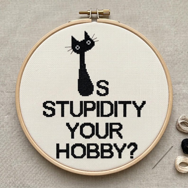 Pu&ograve; includere: Un telaio da ricamo circolare in legno contenente tessuto bianco con un disegno a punto croce nero raffigurante la sagoma di un gatto e il testo IS STUPIDITY YOUR HOBBY? Il disegno &egrave; centrato sul tessuto, teso da una vite in metallo.