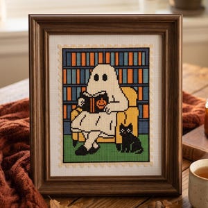May include: Cross-stitch art in a wooden frame. The artwork depicts a ghost reading a book in a yellow armchair, with a black cat at its feet. A bookshelf is in the background. The color palette includes white, orange, blue, green, and black.