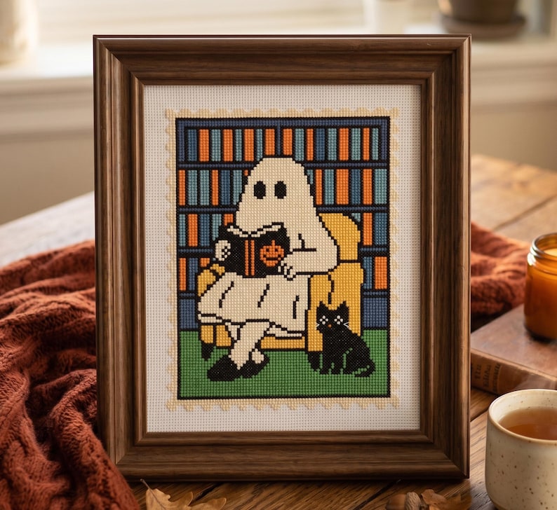 K&ouml;nnte beinhalten: Kreuzstich-Kunstwerk in einem Holzrahmen. Das Kunstwerk zeigt ein Gespenst, das in einem gelben Sessel ein Buch liest, mit einer schwarzen Katze zu seinen F&uuml;&szlig;en. Im Hintergrund ist ein B&uuml;cherregal zu sehen. Die Farbpalette umfasst Wei&szlig;, Orange, Blau, Gr&uuml;n und Schwarz.