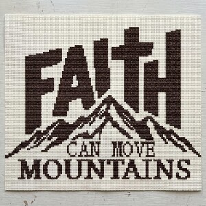 May include: A square cross-stitch embroidery piece on off-white Aida cloth featuring the dark brown text FAITH positioned above a graphic of a mountain range, with the text CAN MOVE MOUNTAINS stitched in dark brown below the mountain design.