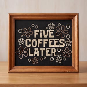May include: A cross-stitch embroidery in a wooden frame featuring the text FIVE COFFEES LATER... in cream-colored thread against a black background, surrounded by small cream-colored flower and circle motifs stitched in a grid pattern.
