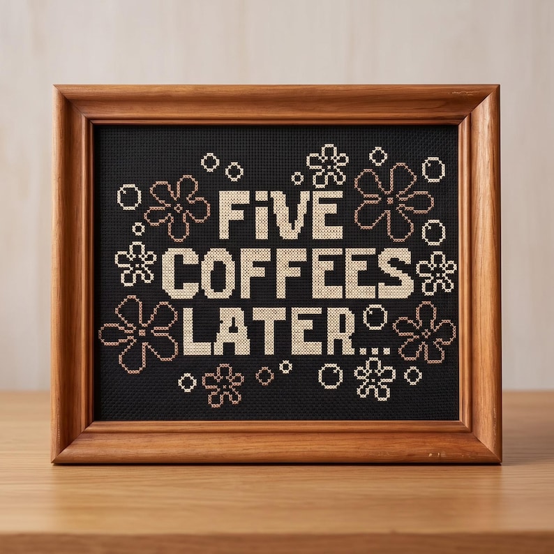May include: A cross-stitch embroidery in a wooden frame featuring the text FIVE COFFEES LATER... in cream-colored thread against a black background, surrounded by small cream-colored flower and circle motifs stitched in a grid pattern.