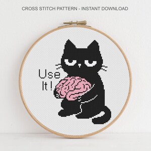K&ouml;nnte beinhalten: Ein Kreuzstichmuster in einem h&ouml;lzernen Stickrahmen mit einer schwarzen Katze mit m&uuml;rrischem Gesichtsausdruck, die ein rosa Gehirn h&auml;lt. Der Text "Use It!" steht links neben der Katze. Der obere Text lautet "CROSS STITCH PATTERN - INSTANT DOWNLOAD".