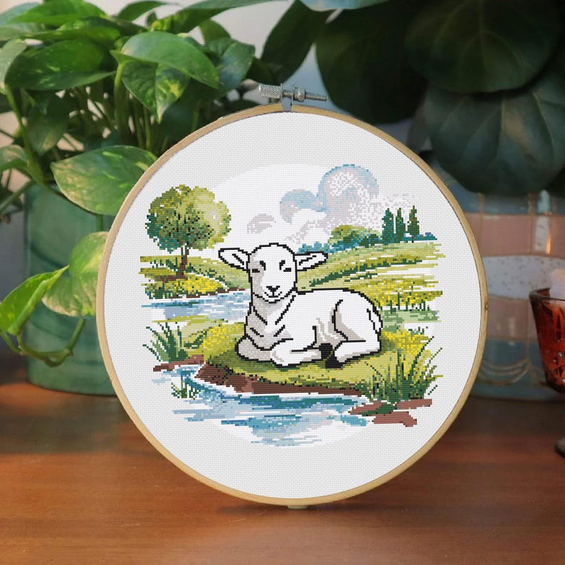 May include: A cross-stitch embroidery hoop featuring a lamb resting near a stream. The scene includes a tree, grassy banks, and a cloudy sky, all rendered in soft blues, greens, and whites. The embroidery is set in a wooden hoop.