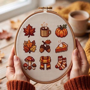 Könnte beinhalten: Ein runder Holzstickrahmen mit einem Kreuzstichmuster auf weißem Stoff. Das Design zeigt neun herbstliche Symbole: ein Ahornblatt, eine Tasse Kakao, einen Kürbis, Eichenblätter, Eicheln, ein Stück Kuchen, Pilze, einen Schal und Socken.