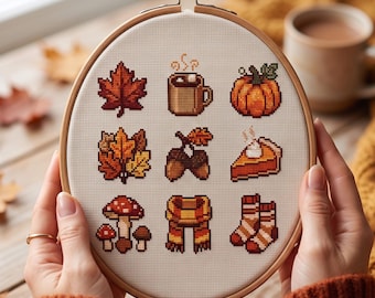 May include: A round wooden embroidery hoop containing a cross-stitch pattern on white fabric. The design features nine autumn-themed icons: a maple leaf, a mug of cocoa, a pumpkin, oak leaves, acorns, a slice of pie, mushrooms, a scarf, and socks.
