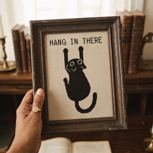 K&ouml;nnte beinhalten: Ein gerahmtes Kreuzstich-Stickbild mit der Silhouette einer schwarzen Katze, die oben h&auml;ngt, und dem Text HANG IN THERE in schwarzem Garn auf beigem Stoffhintergrund, eingefasst in einen dunkelbraunen, abgenutzten Holzrahmen.