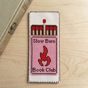 May include: A rectangular cross-stitch bookmark on white fabric featuring a matchbox design. The top shows brown matches with red tips. The pink box below displays the text Slow Burn Book Club in dark brown letters with a red flame graphic.