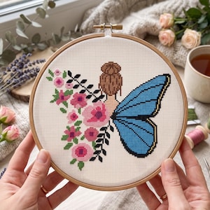 Peut inclure: Un tambour &agrave; broder en bois circulaire contenant un motif au point de croix sur tissu blanc. Le dessin repr&eacute;sente une personne avec un chignon brun, une grande aile de papillon bleue et un arrangement vertical de fleurs roses et de feuilles noires.