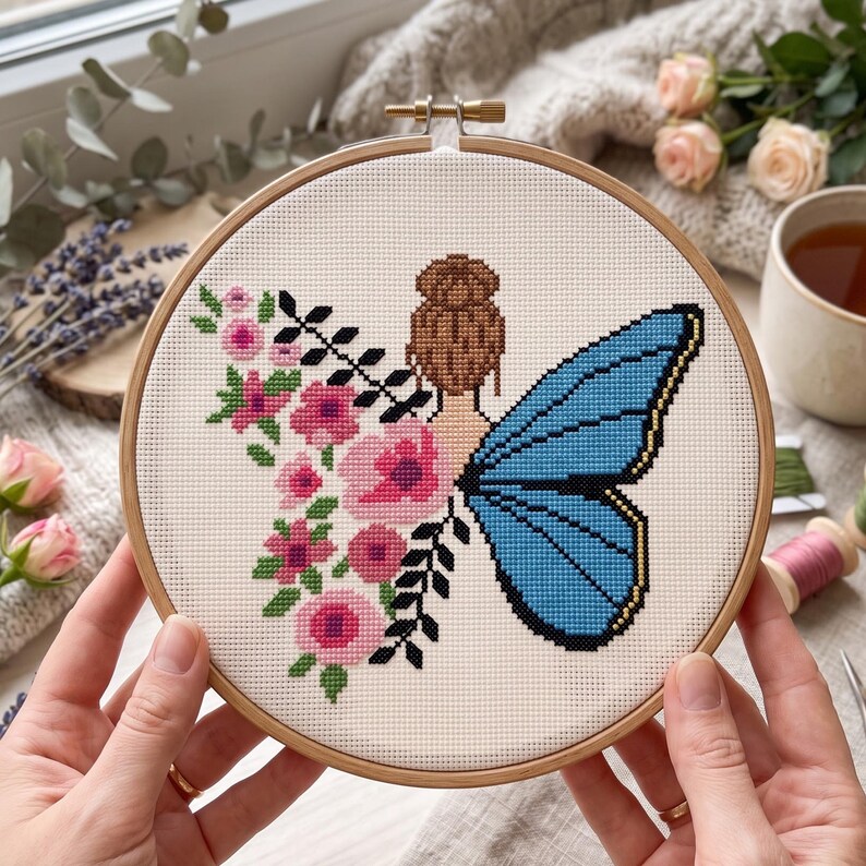 Peut inclure: Un tambour &agrave; broder en bois circulaire contenant un motif au point de croix sur tissu blanc. Le dessin repr&eacute;sente une personne avec un chignon brun, une grande aile de papillon bleue et un arrangement vertical de fleurs roses et de feuilles noires.