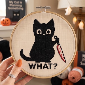 May include: A circular wooden embroidery hoop containing a cross-stitch design on cream fabric. The design features a black cat with large white eyes holding a knife with a red and white blade. The word WHAT? is stitched in black below the cat.