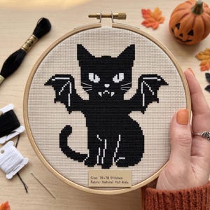 Puede incluir: Un proyecto de punto de cruz en un bastidor de madera que muestra un gato negro con alas de murci&eacute;lago, ojos blancos y colmillos sobre tela Aida natural. Una etiqueta en el bastidor indica: Size: 78x78 Stitches, Fabric: Natural 14ct Aida. Hilo incluido.