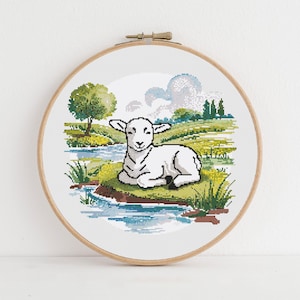 May include: A cross-stitch embroidery of a lamb resting near a stream. The artwork is set in a wooden hoop and features a pastoral scene with a tree, grass, and a blue sky with clouds. The lamb is white with black outlines.