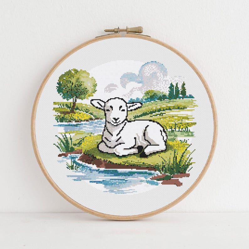 May include: A cross-stitch embroidery of a lamb resting near a stream. The artwork is set in a wooden hoop and features a pastoral scene with a tree, grass, and a blue sky with clouds. The lamb is white with black outlines.