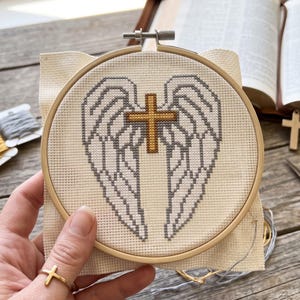 May include: A circular wooden embroidery hoop containing a cross-stitch pattern on cream-colored fabric. The design features a pair of symmetrical grey-outlined angel wings with a central gold-colored cross positioned between the wings.
