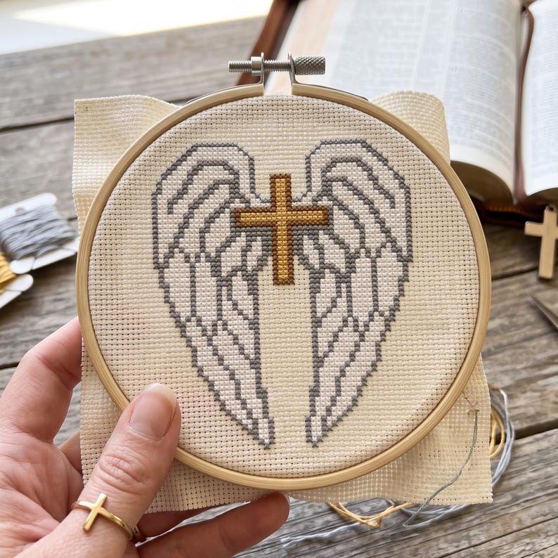 May include: A circular wooden embroidery hoop containing a cross-stitch pattern on cream-colored fabric. The design features a pair of symmetrical grey-outlined angel wings with a central gold-colored cross positioned between the wings.