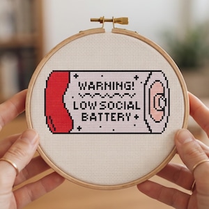 May include: A circular wooden embroidery hoop containing white fabric with a cross-stitch design of a battery icon. The design features a red end, a pink terminal, and black text that reads WARNING! LOW SOCIAL BATTERY.